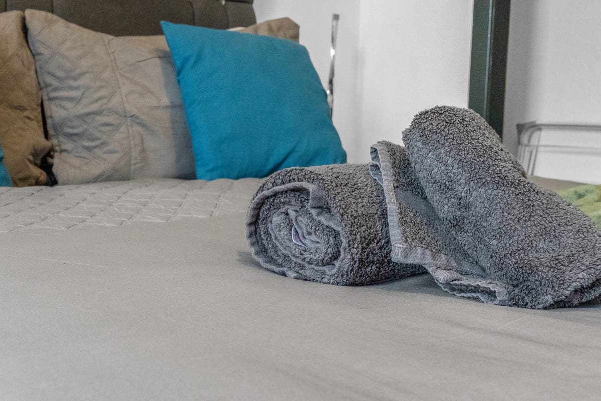 A bed made up with white linen, grey and teal cushions, and rolled towels set out at the foot.
