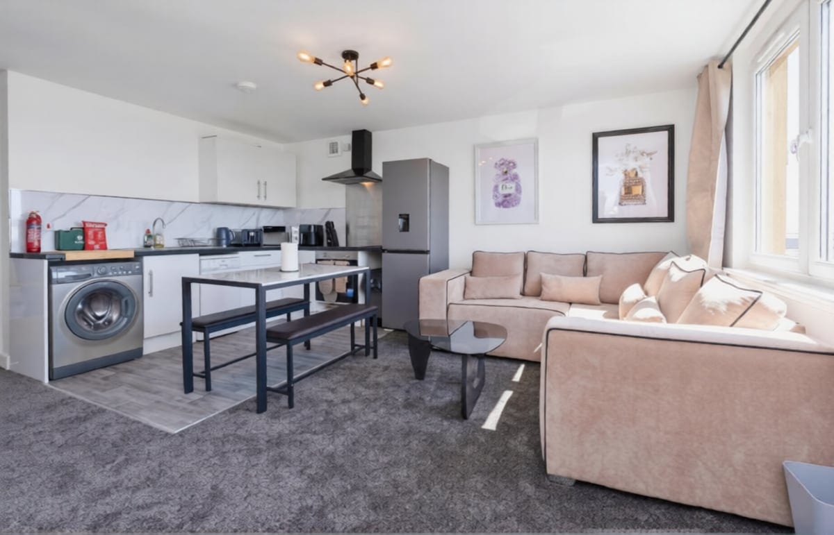Open-plan kitchen and living room with white units, a washing machine, a breakfast bar, and a beige corner sofa.