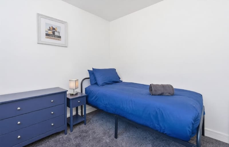 Single bedroom with a blue chest of drawers, a bedside lamp, and folded towels on the bed.