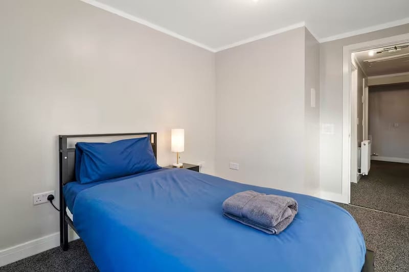 Bedroom with a bed in blue linen, a bedside lamp, and the door open to the hallway.