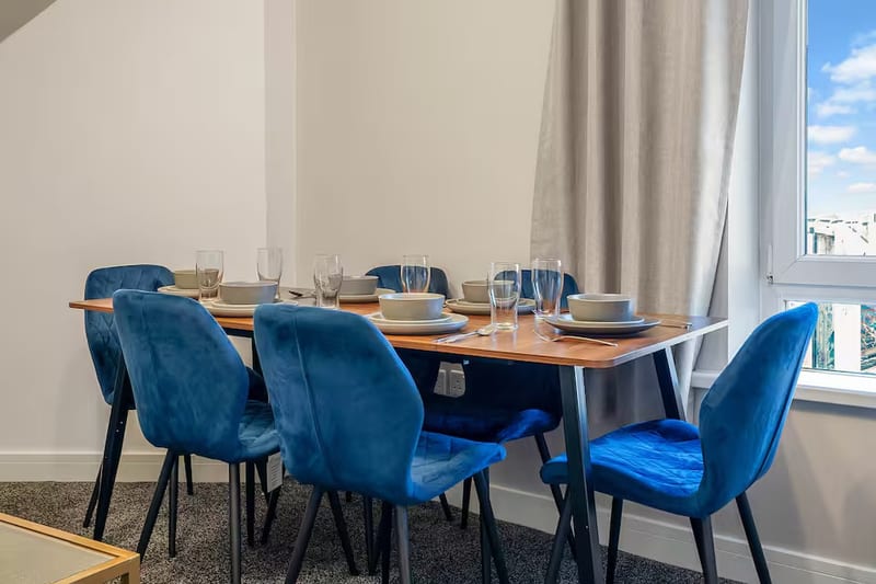 Dining table laid for six with grey bowls and glasses, under a window with grey curtains.