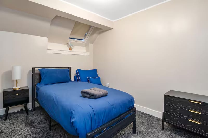 Double bedroom with blue linen, a black bedside table and lamp, and a chest of drawers under a skylight.