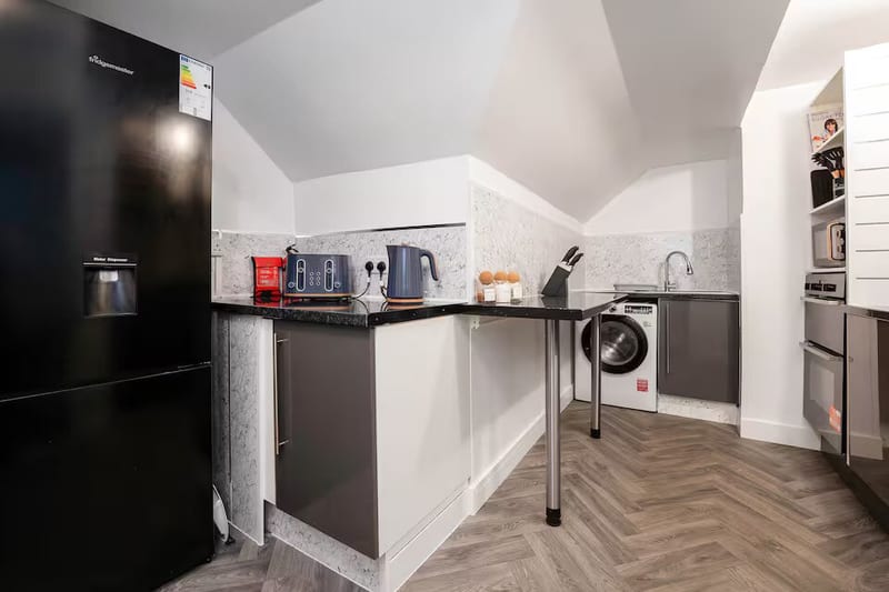 Kitchen with dark grey units, a black fridge freezer, a washing machine and a herringbone wood floor.
