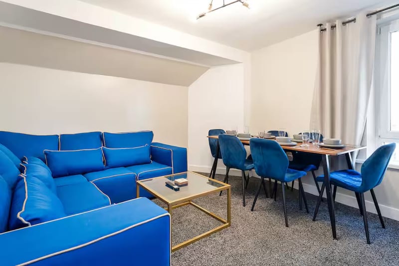Living room with a blue corner sofa and a gold-framed coffee table, opening onto a dining table with six blue velvet chairs.