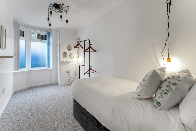 Bedroom with a bed dressed in grey, a bay window, and pendant lights on the wall.