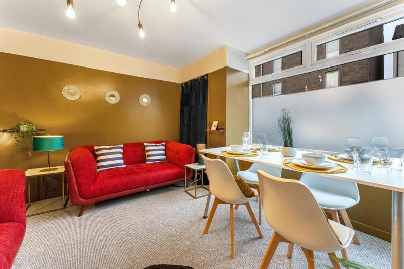 Living and dining room with ochre walls, a red sofa, and a dining table with four white chairs by the window.