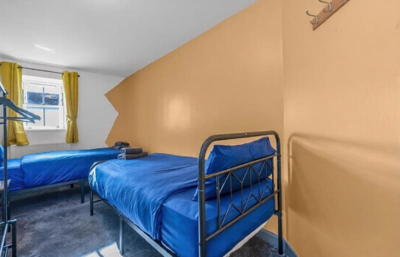 Bedroom with two single beds in blue linen under a sloped ceiling, with yellow curtains at the window.