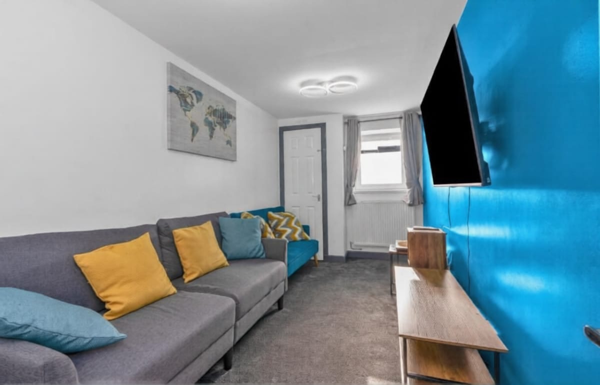 Living room with a grey corner sofa, mustard and teal cushions, and a wall-mounted television on a blue feature wall.