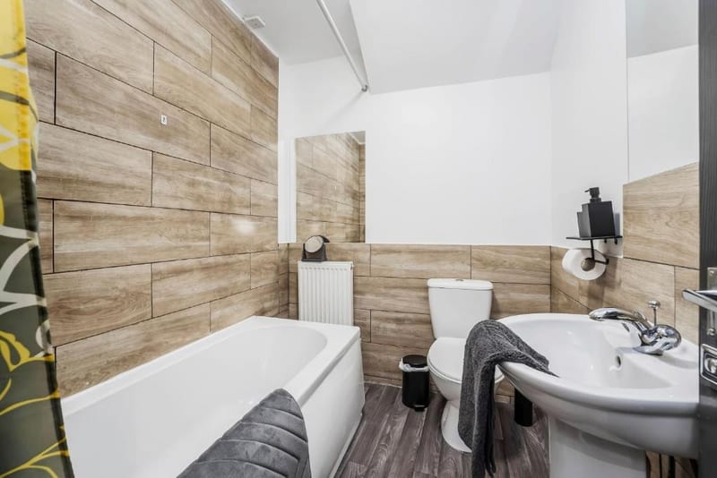 Bathroom with a bath, wood-effect tiled walls, a toilet and a pedestal basin.