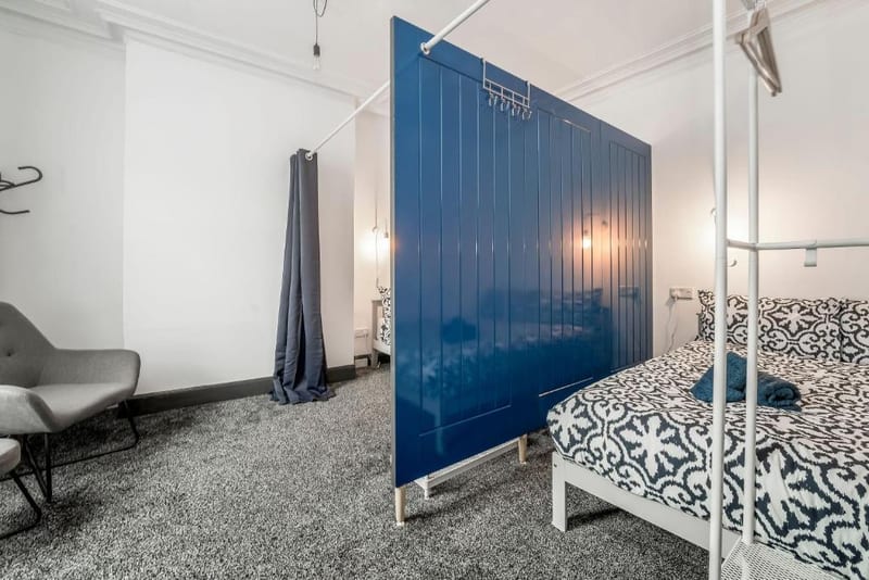 Double bed with a black and white patterned cover behind a tall blue panelled divider, with an armchair to one side.