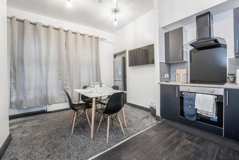 Dining table for four on a grey carpet beside the kitchen, with long pale curtains behind and the oven to the right.