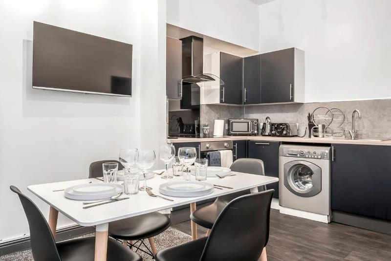 Dining table laid for four in front of a kitchen with dark grey units and a washing machine, with a wall-mounted television beside it.