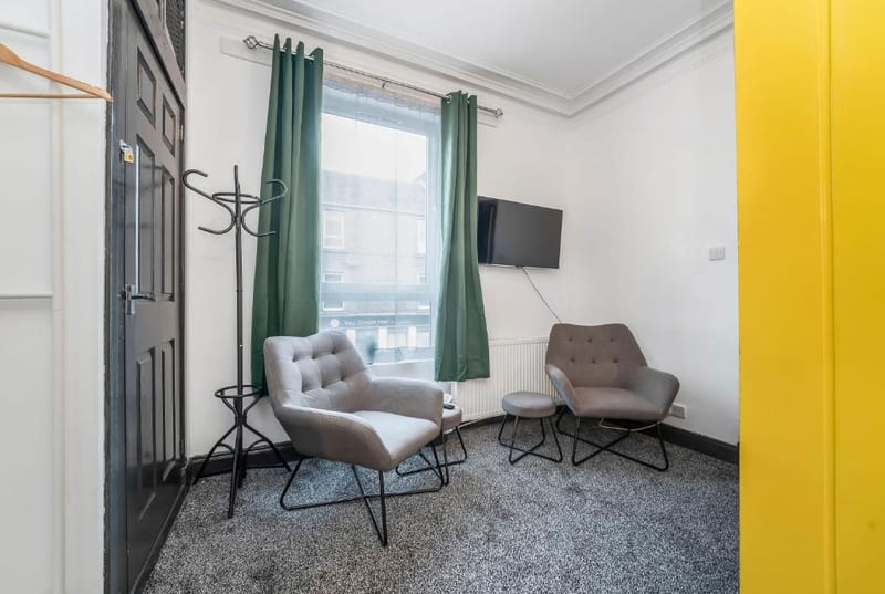 Sitting area with two grey armchairs and a footstool by a window with green curtains, and a wall-mounted television.