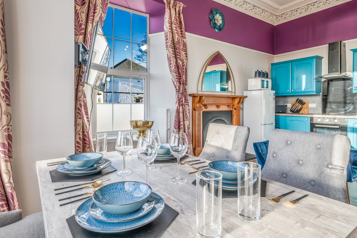 Dining table laid for four with blue plates and wine glasses, in a room with a wooden fireplace and a tall window.