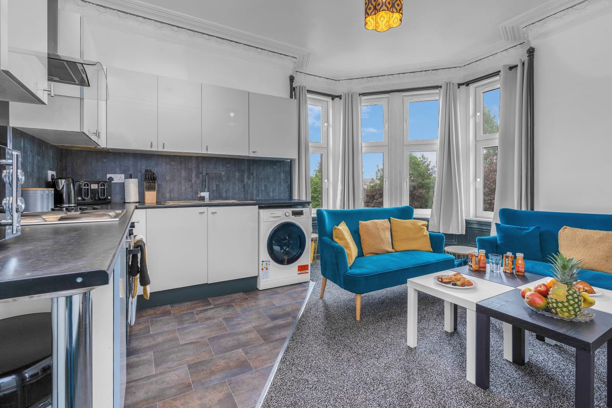 Open-plan kitchen and sitting room with a bay window, a teal two-seat sofa and armchair, and a washing machine built into the run of units.