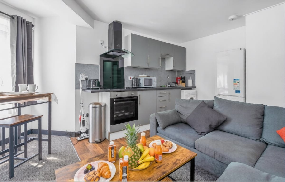Open-plan kitchen and living room with grey units, a grey sofa, and breakfast laid out on a wooden coffee table.