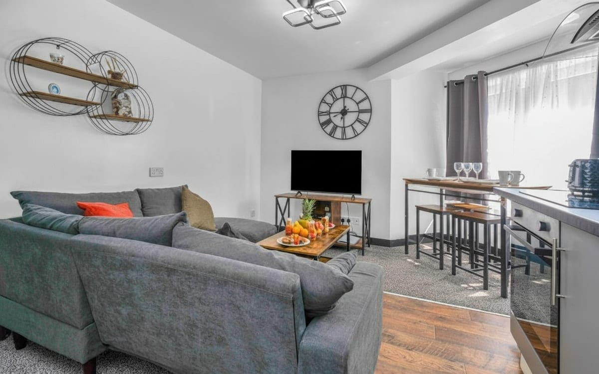 Living room with a grey corner sofa, a wall-mounted television and a large round clock, with a breakfast bar behind.