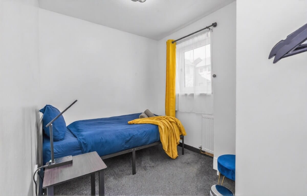 Single bedroom with a blue bed, a yellow throw, and a desk under the window.