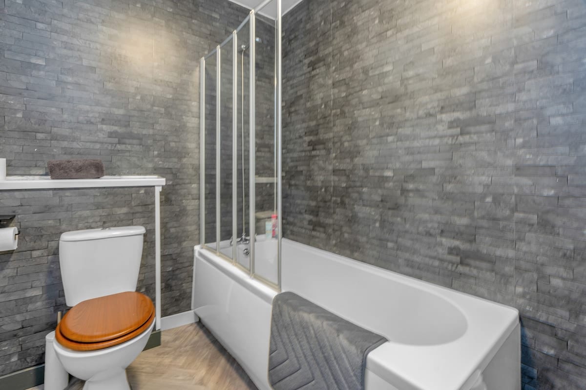 Bathroom with a bath and shower screen, a slate-effect tiled wall, and a wooden toilet seat.