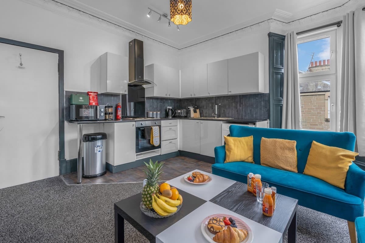 Kitchen and living room with white units, a teal sofa with mustard cushions, and a low table set for breakfast.