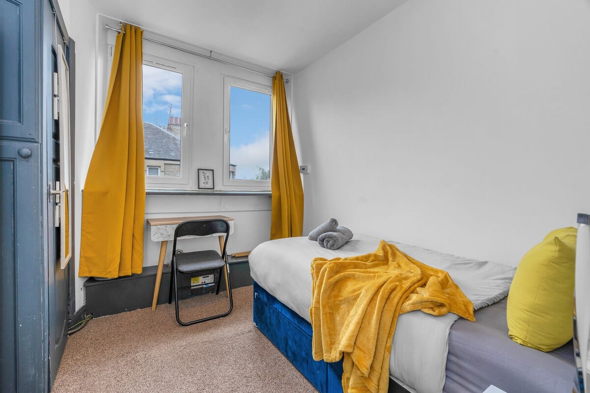 Single bedroom with a mustard throw, a folding desk, and yellow curtains at the window.