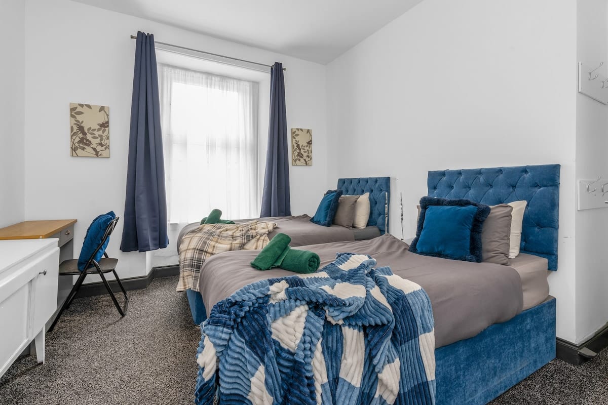 Twin room with two beds, blue buttoned headboards and folded towels, beside a window with grey curtains.