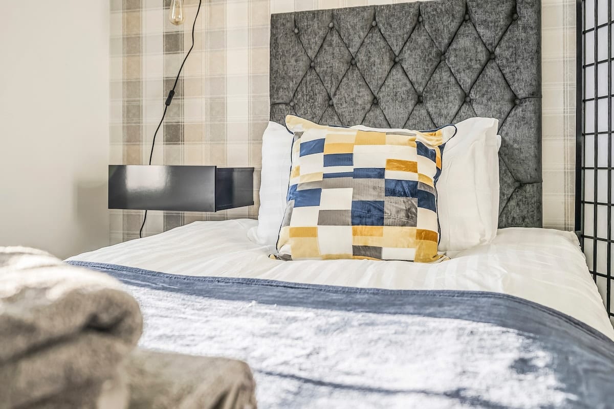 Bed with a grey buttoned headboard, a checked cushion, and a velvet throw.