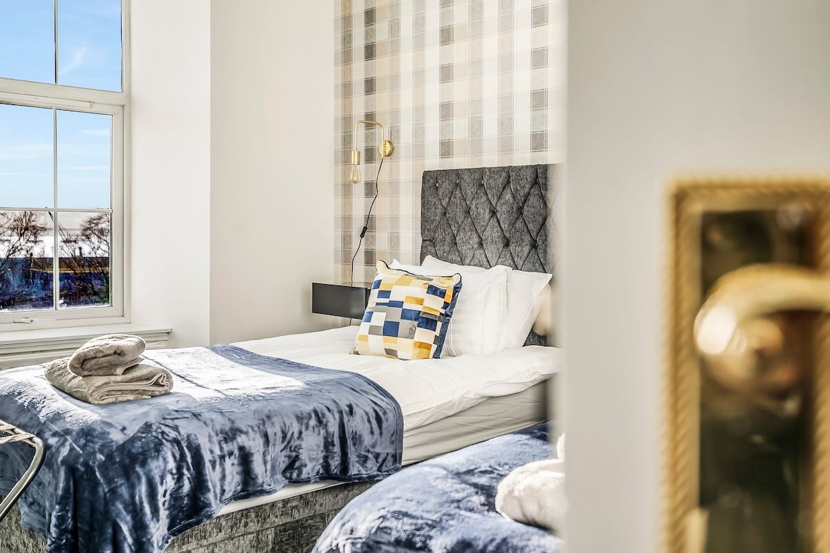Bedroom with a grey buttoned headboard, folded towels, and a window looking towards the water.