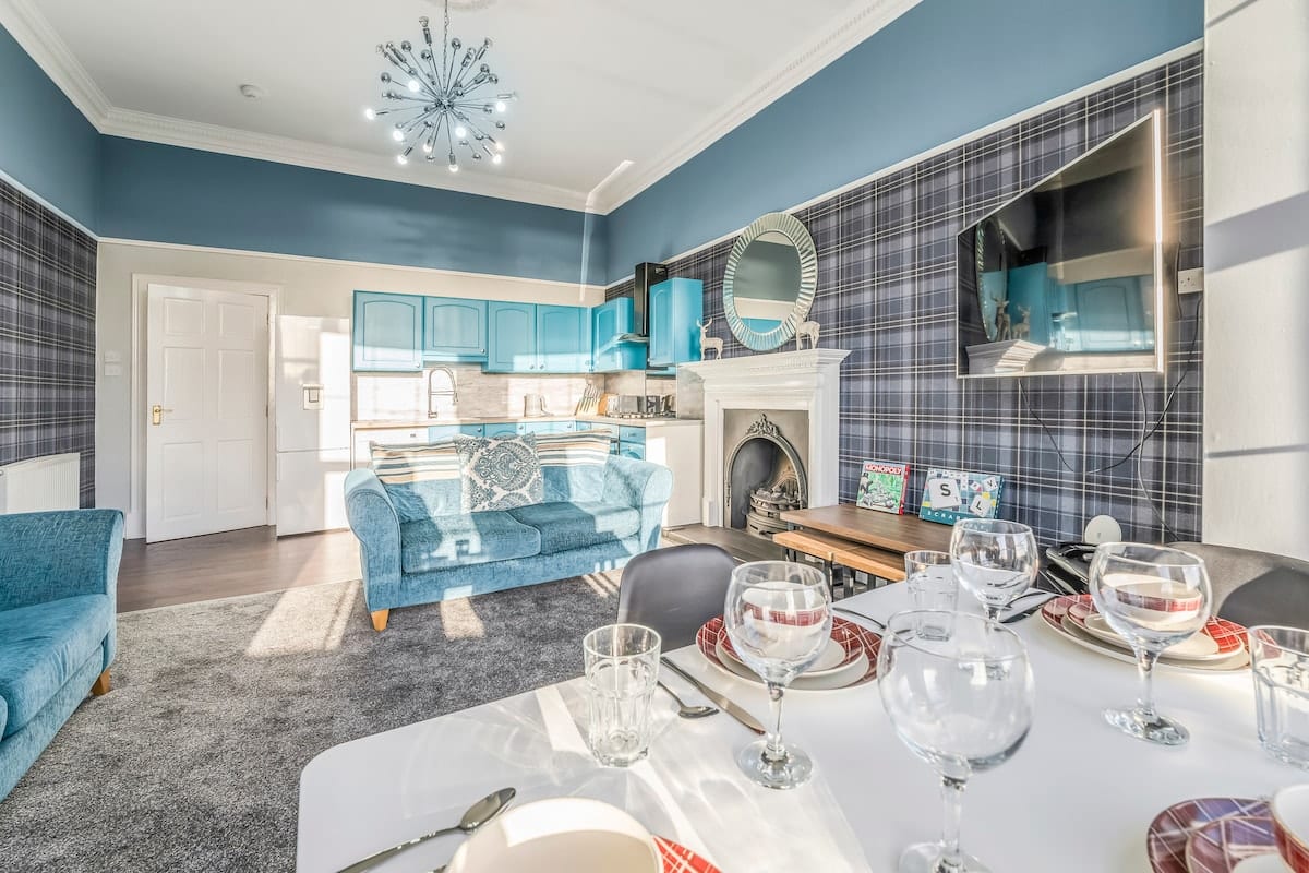Sitting and dining room with a tartan wall, a teal kitchen behind, and the table laid with wine glasses.