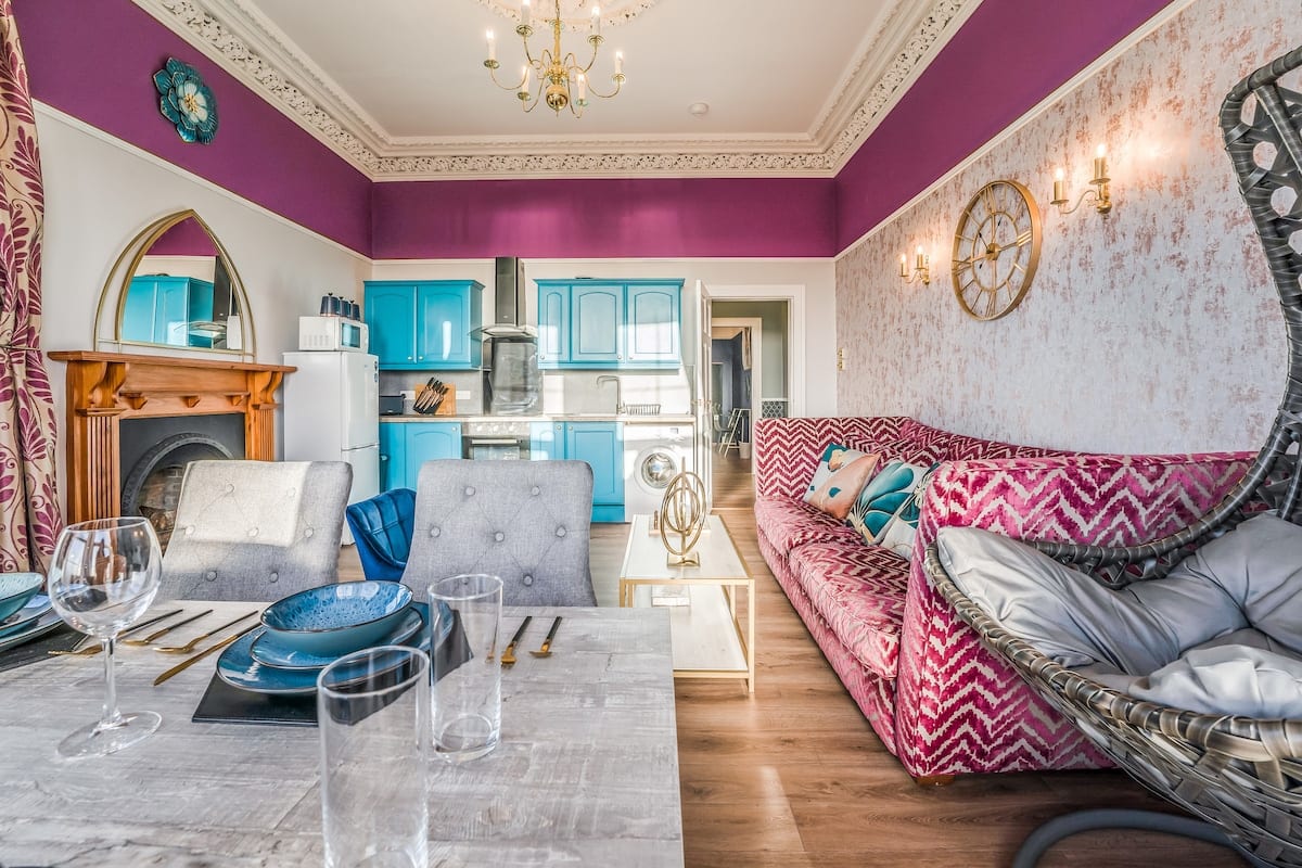 Dining table laid with blue plates and wine glasses, with a wooden fireplace, a teal kitchen, and a patterned pink sofa.
