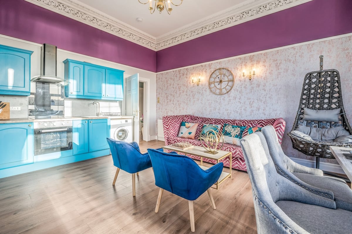 Teal kitchen with an oven and extractor, seen across the sitting room from the blue armchairs.