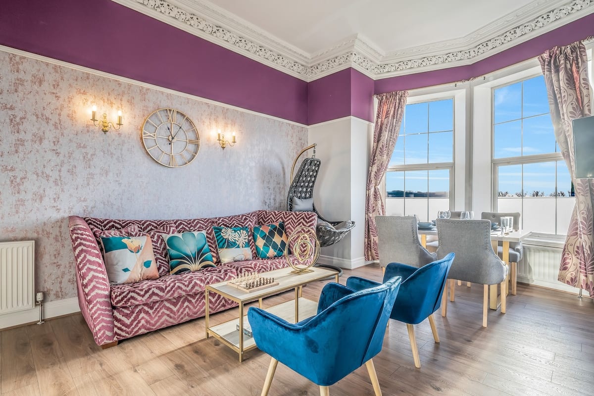 Sitting room with a patterned sofa, blue velvet armchairs, and a bay window over the water.