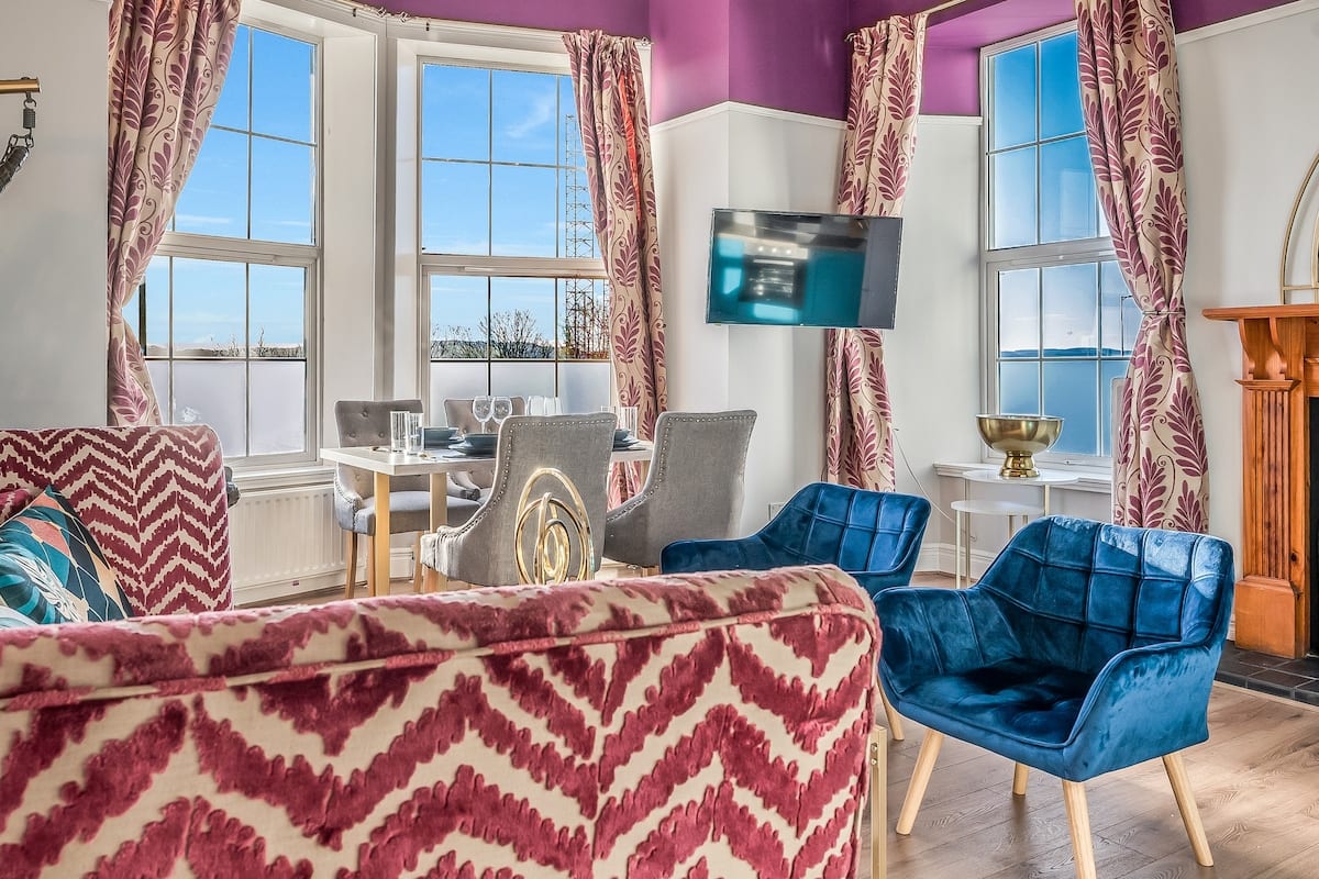 Sitting room with a patterned pink sofa, blue velvet chairs, and tall windows onto the River Tay.