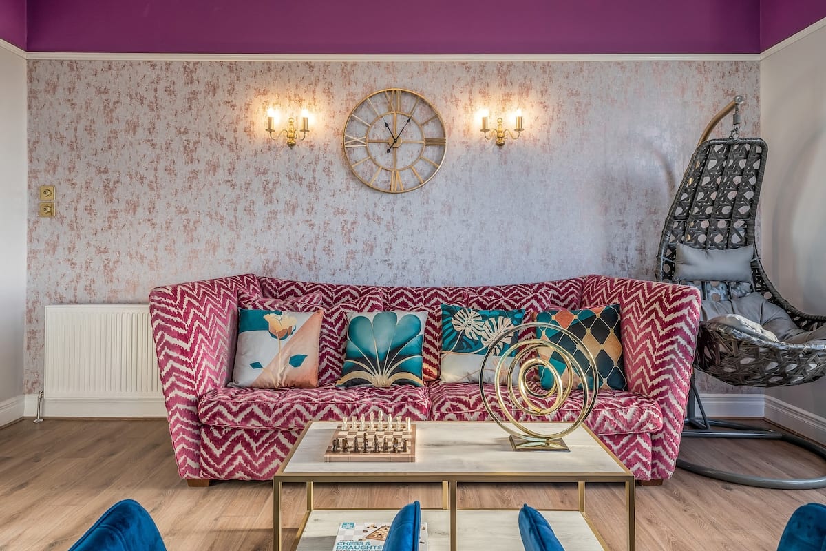 Patterned pink sofa below a starburst clock and wall lights, with a hanging chair in the corner.