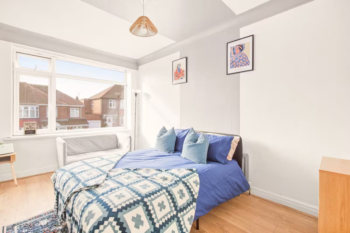 Bedroom with a double bed in blue linen, a padded bench at its foot and a bay window over the rooftops.