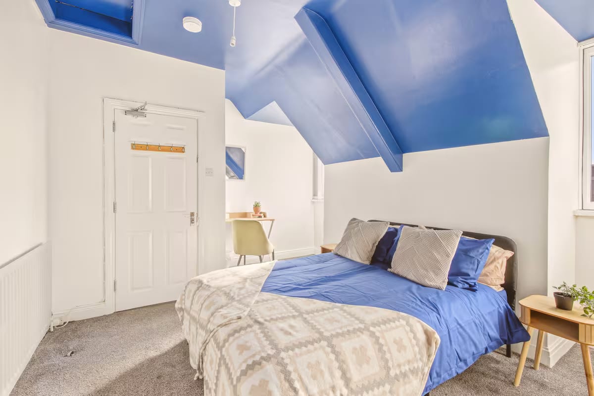 Bedroom under a blue-painted sloped ceiling, with a double bed in blue linen and a small desk and chair by the window.