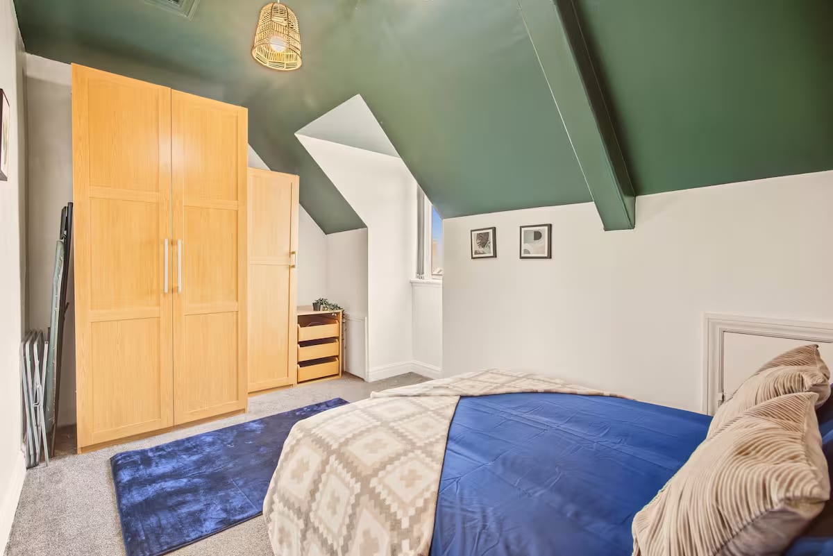 Bedroom under a green-painted sloped ceiling, with a double bed in blue linen, a tall pine wardrobe and a chest of drawers.
