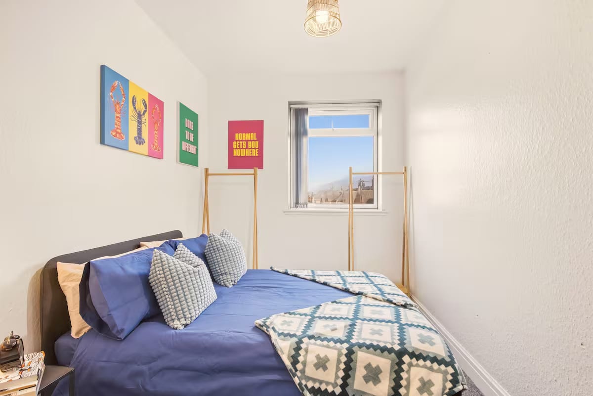 Bedroom with a double bed in blue linen, bright framed prints above the bed and a window at the far end.