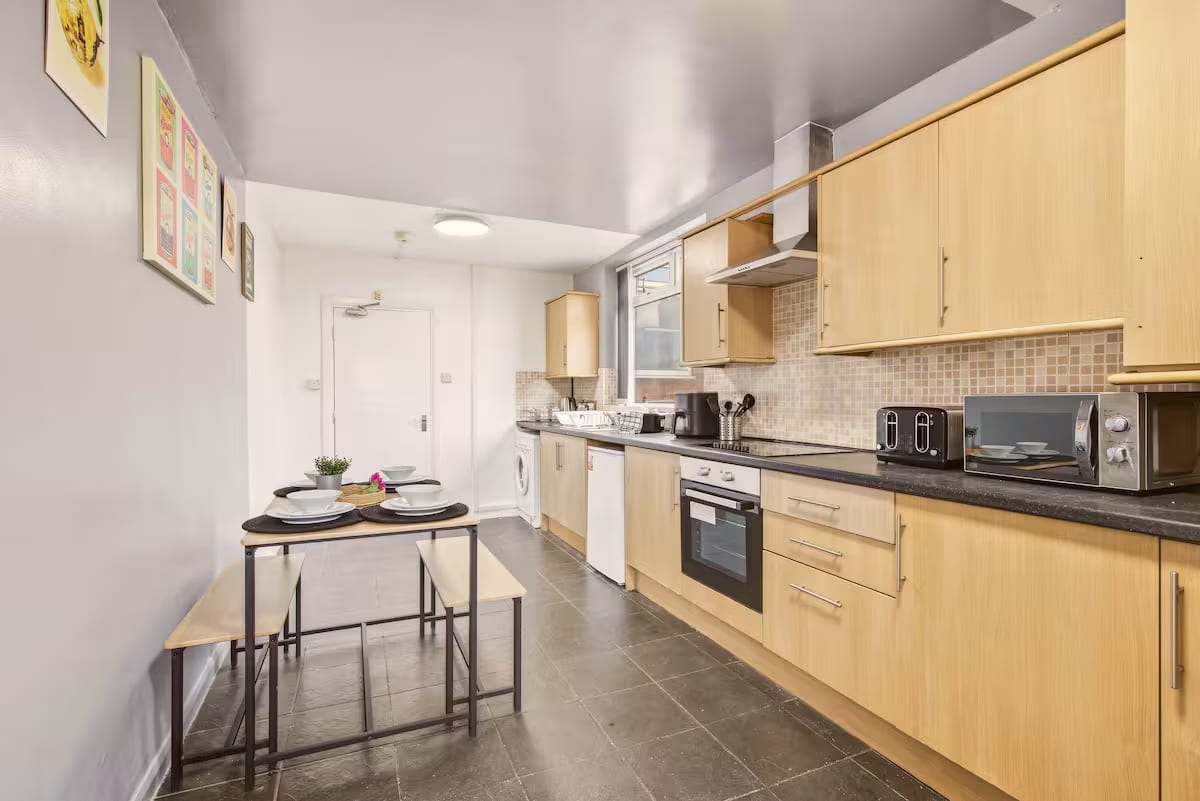 Galley kitchen with wood units, an oven, a microwave and a toaster along a dark worktop, and a small dining table with two benches.