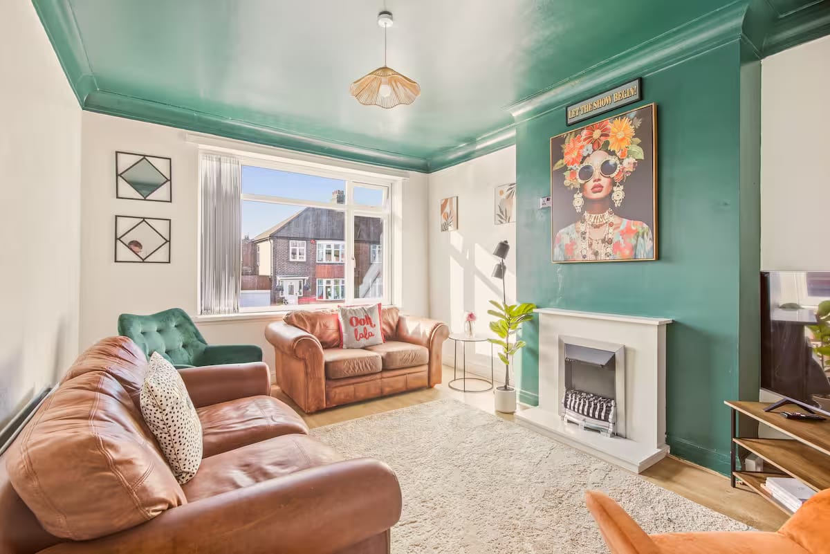Living room with a green ceiling and chimney breast, two brown leather sofas, a green velvet armchair and a bay window onto the street.