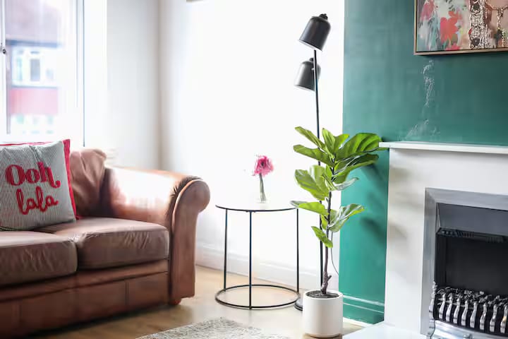 Corner of the living room: the arm of a leather sofa, a black floor lamp, a plant and a small round table beside the fireplace.